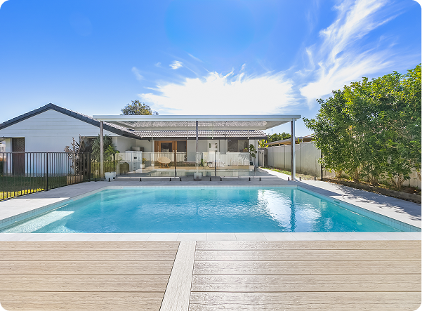 Modern backyard pool with a shaded patio and lush greenery under a blue sky.
